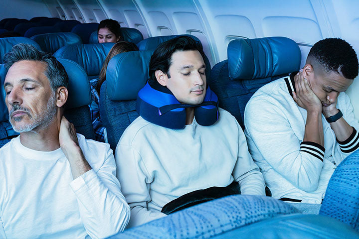 Three men sitting in airplane seats, two with neck pillows, in a cabin setting.