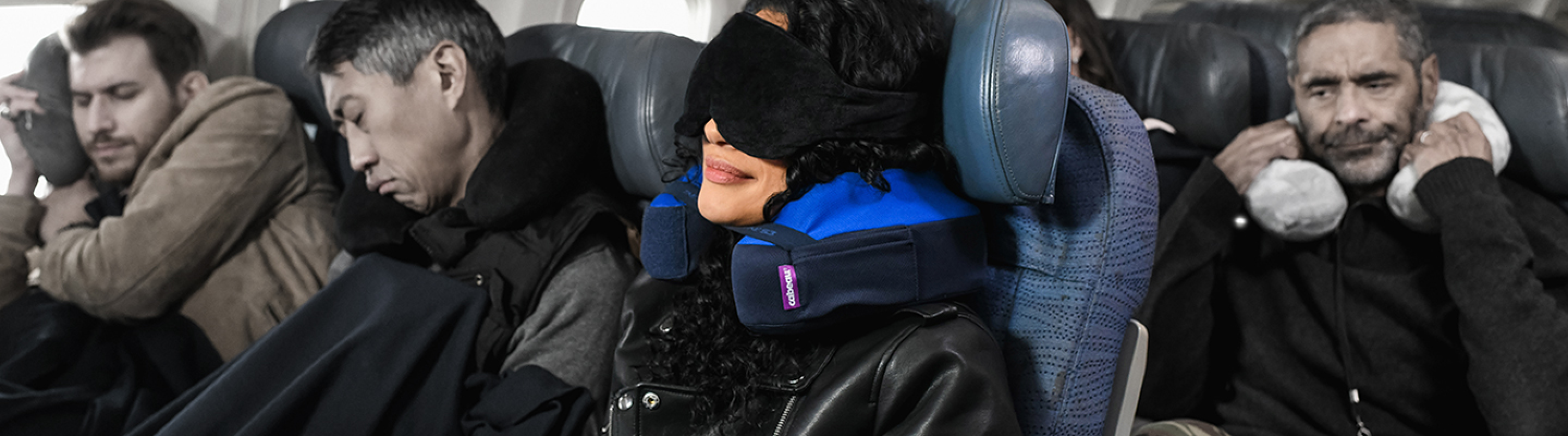 Traveler wearing TNE S3 neck pillow and sleep mask comfortably on airplane while other passengers look uncomfortable around her